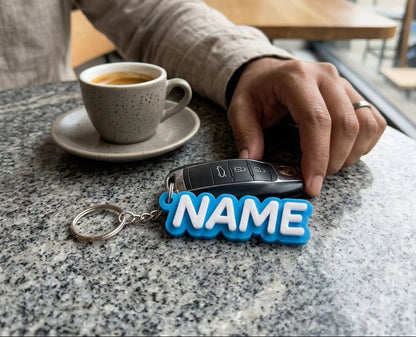 Person holding a keychain with 'NAME' on it next to a cup of coffee in a cafe.